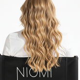Long, wavy blonde hair shown from the back on a model wearing a white shirt and a black chair with the brand name NIOMI visible.