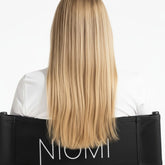 Long, straight blonde hair shown from the back on a model wearing a white top and sitting in a black chair with the brand name NIOMI visible.