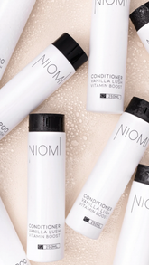 Several white NIOMI Hair Care bottles with black caps are arranged on a light beige textured surface with a water droplet effect. The visible text identifies the product as Vanilla Lush Vitamin Boost Conditioner.